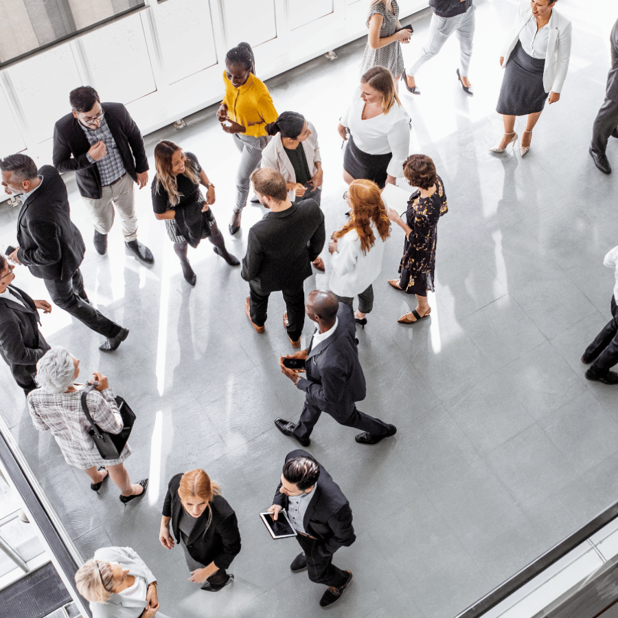 A bird's-eye view of business professionals in formal attire mingling and networking in a modern office lobby or conference center with polished floors and white architectural elements.