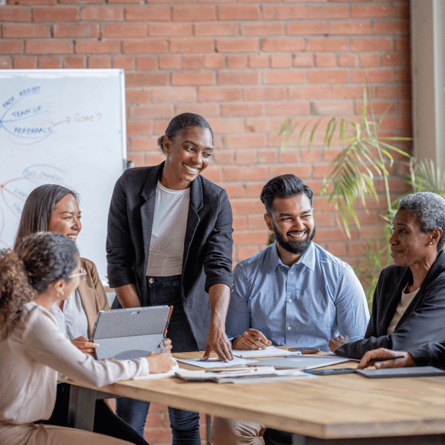 A diverse group of five professionals engaged in a collaborative meeting around a wooden conference table, with a smiling woman in a dark blazer standing and presenting while others are seated and attentively listening. The modern office setting features exposed brick walls, a whiteboard, and a plant in the background.