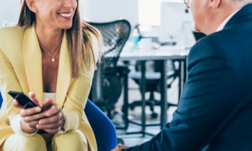 A woman in a bright yellow blazer smiles while holding a smartphone during a business conversation with an older man in glasses and a dark suit in a modern office setting.