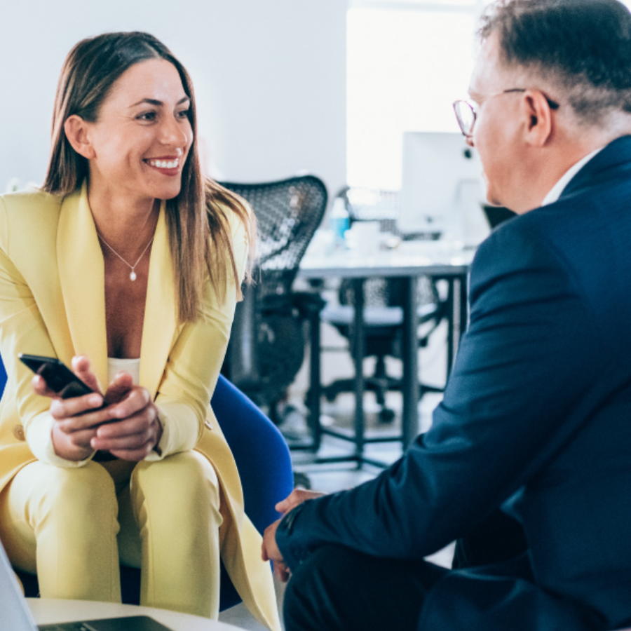 A woman in a bright yellow blazer smiles while holding a smartphone during a business conversation with an older man in glasses and a dark suit in a modern office setting.
