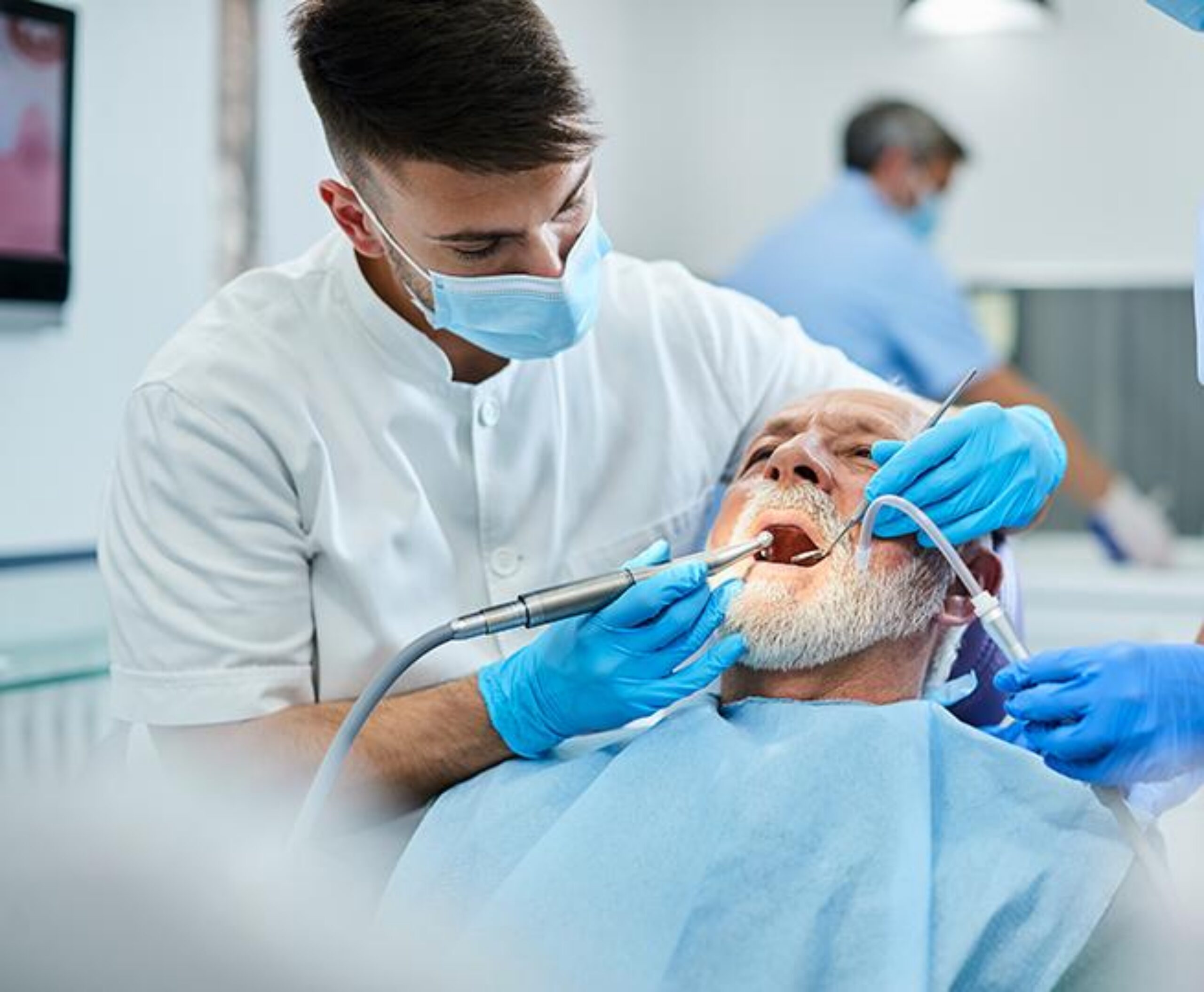 A dentist wearing a surgical mask and blue gloves examines an elderly patient's mouth using dental instruments in a modern dental office, with another medical professional visible in the background.