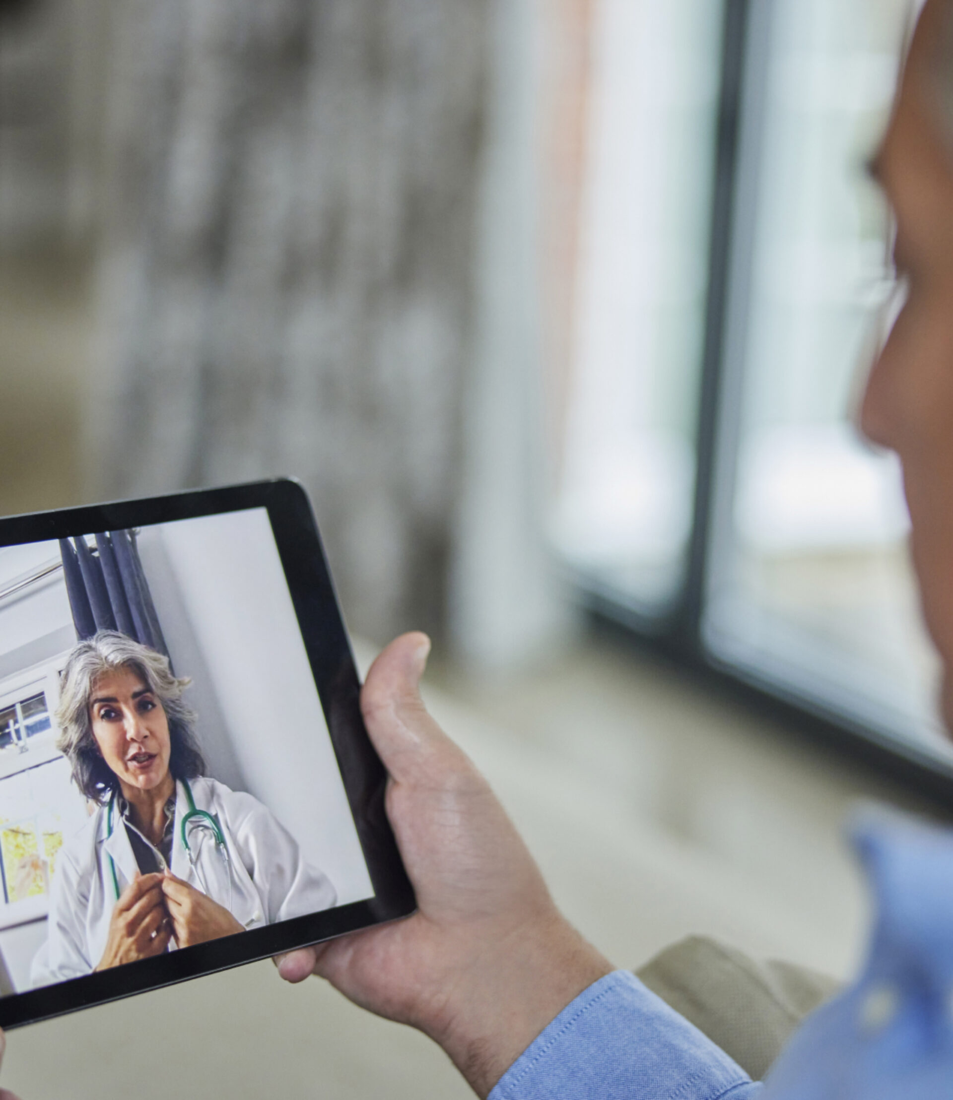 A person holds a tablet displaying a video call with a healthcare professional wearing a white coat and stethoscope, illustrating a telemedicine consultation.