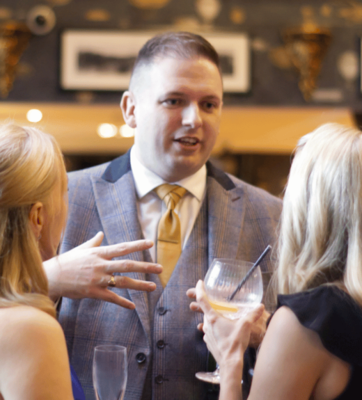A man in a gray suit and yellow tie speaks with two blonde women at what appears to be a social gathering or networking event, with one woman holding a wine glass.