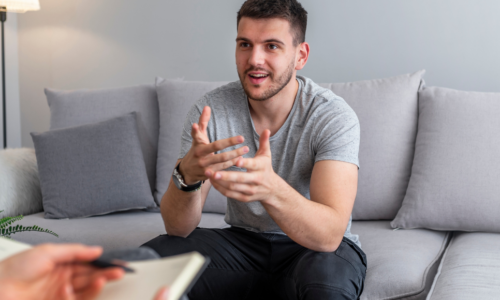 A young man in a gray t-shirt sits on a light gray couch, gesturing with his hands while speaking to someone whose hands and notebook are visible in the foreground, suggesting a counseling or interview session.