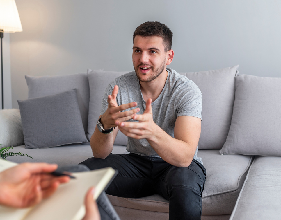 A young man in a gray t-shirt sits on a light gray couch, gesturing with his hands while speaking to someone whose hands and notebook are visible in the foreground, suggesting a counseling or interview session.