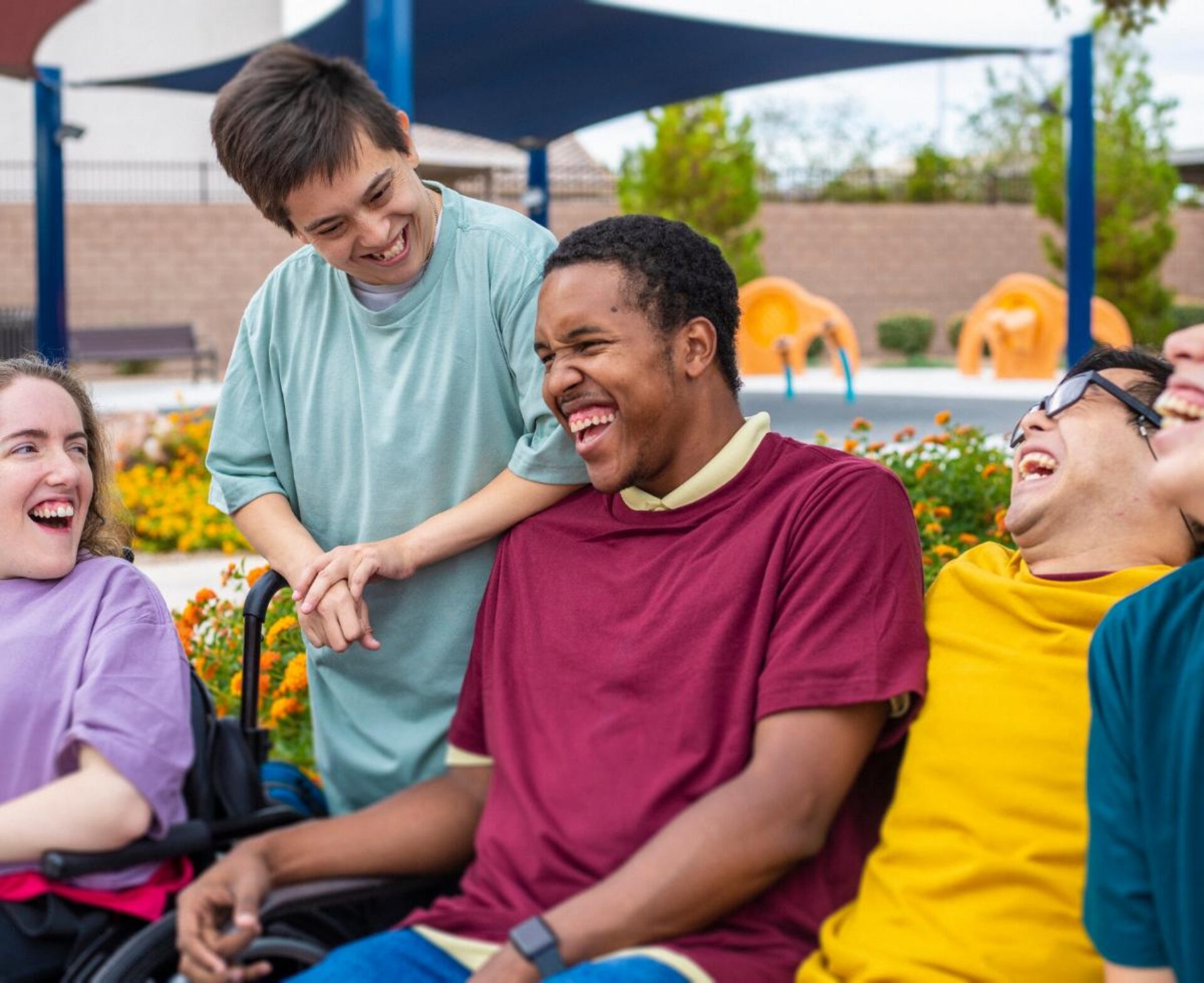 A group of young people with disabilities laughing and socializing together outdoors, with one person standing behind a friend in a wheelchair in a supportive, friendly gesture at what appears to be a recreational facility or park.