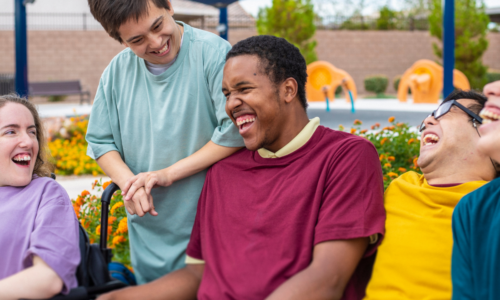 A group of young people with disabilities laughing and socializing together outdoors, with one person standing behind a friend in a wheelchair in a supportive, friendly gesture at what appears to be a recreational facility or park.