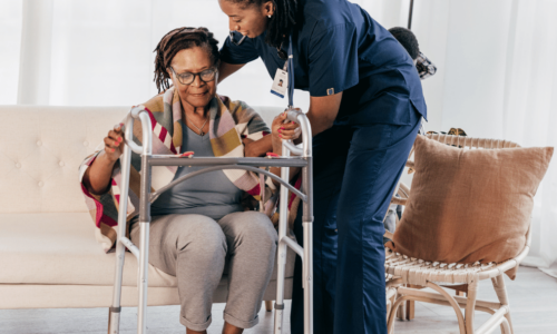 A healthcare worker in blue scrubs assists an elderly woman with glasses who is using a walker in a bright, modern living room setting.
