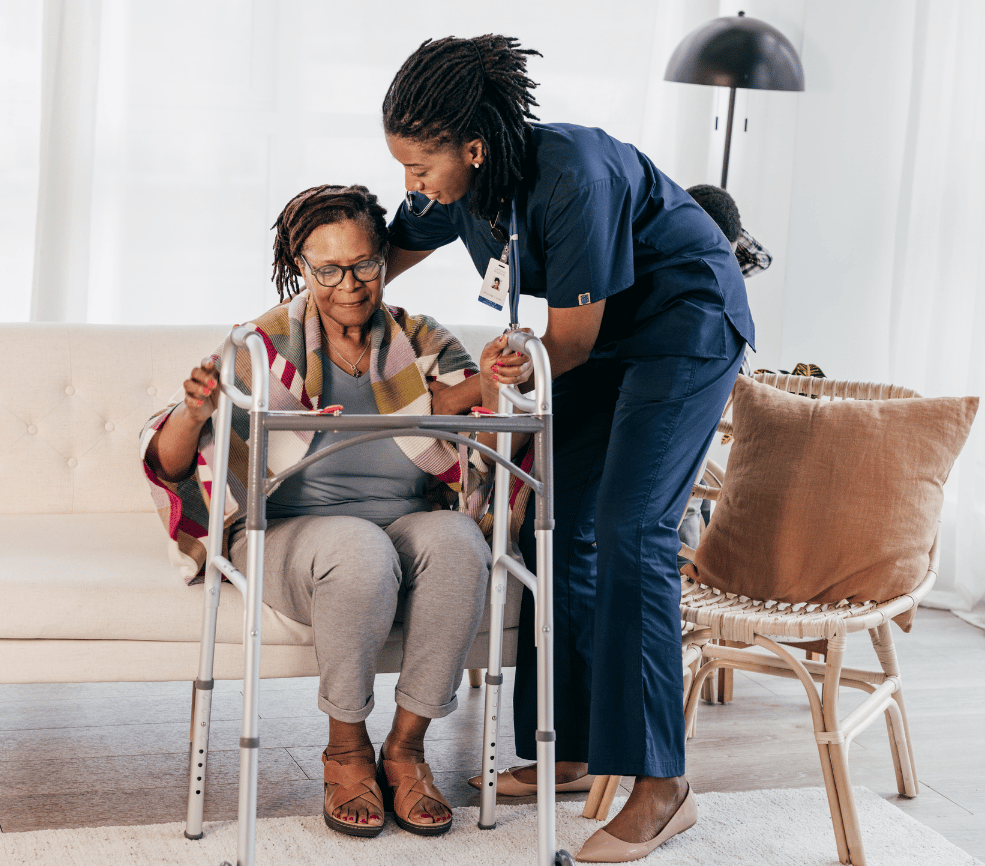A healthcare worker in blue scrubs assists an elderly woman with glasses who is using a walker in a bright, modern living room setting.
