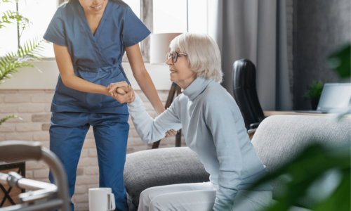 A healthcare worker in blue scrubs helps an elderly woman with white hair and glasses stand up from a gray couch in a bright, modern living room setting.