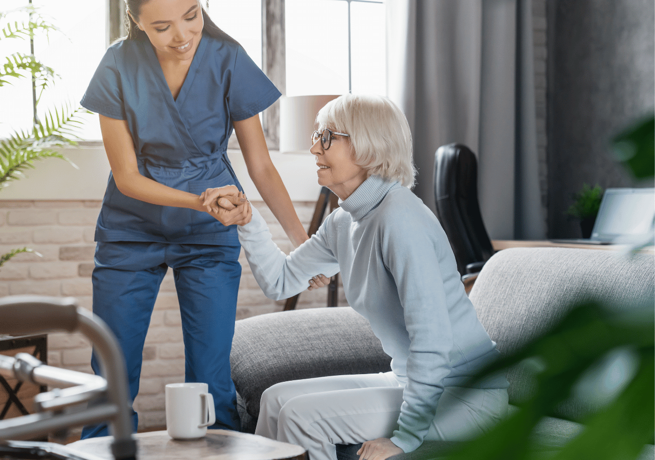 A healthcare worker in blue scrubs helps an elderly woman with white hair and glasses stand up from a gray couch in a bright, modern living room setting.