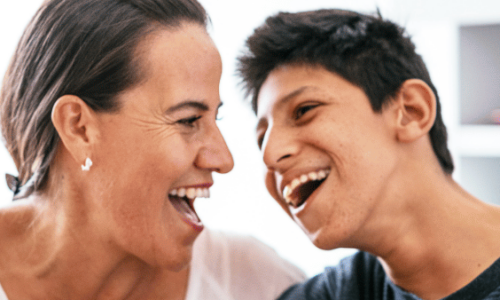 A woman and young man laugh joyfully together in close proximity, appearing to share a warm, happy moment in what looks like a home setting.