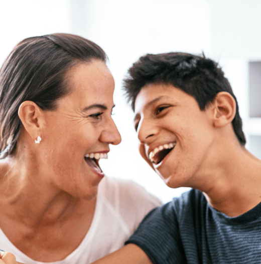 A woman and young man laugh joyfully together in close proximity, appearing to share a warm, happy moment in what looks like a home setting.