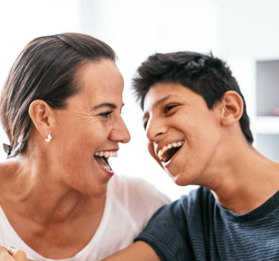 A woman and young man laugh joyfully together in close proximity, appearing to share a warm, happy moment in what looks like a home setting.