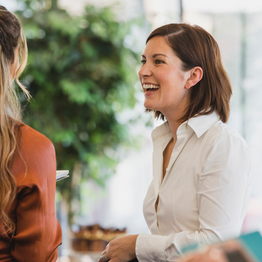 A woman in a white button-up shirt smiles warmly while engaged in conversation with another person in an orange top, sitting in what appears to be a bright, plant-filled indoor space.