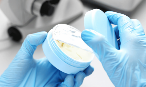 A scientist wearing blue latex gloves examines a petri dish containing yellow bacterial or cellular cultures in a laboratory setting with microscope equipment visible in the blurred background.