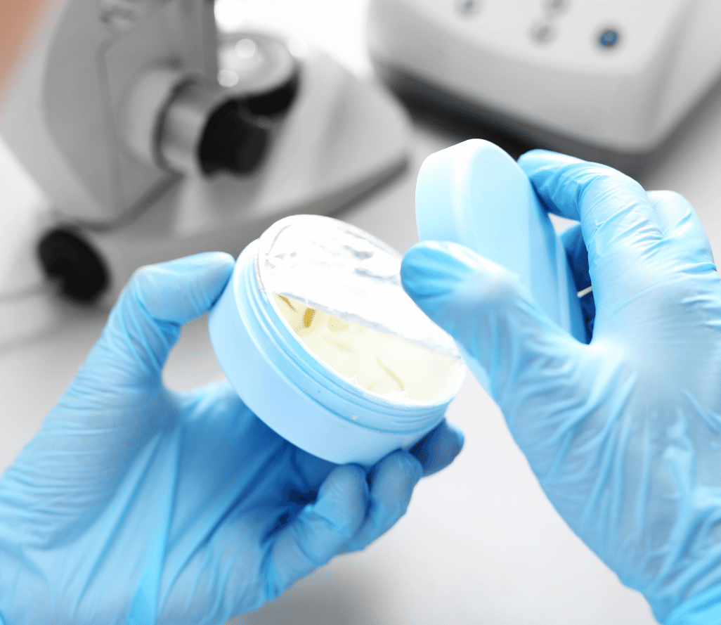 A scientist wearing blue latex gloves examines a petri dish containing yellow bacterial or cellular cultures in a laboratory setting with microscope equipment visible in the blurred background.