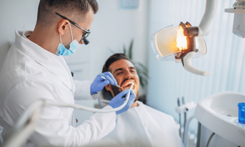 A dentist wearing glasses, a mask, and blue gloves examines a smiling male patient in a modern dental office with professional lighting and equipment.