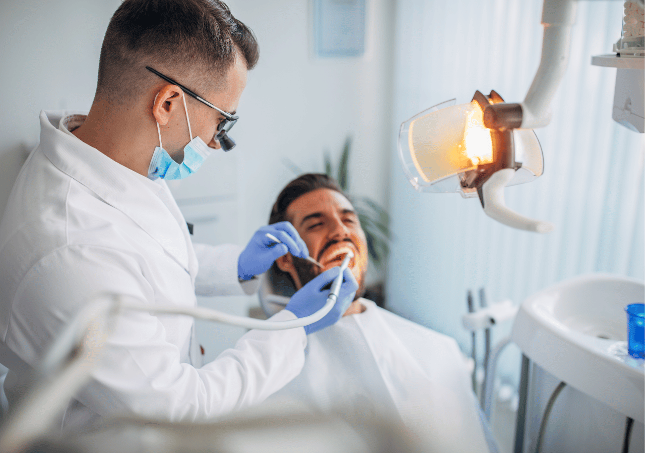 A dentist wearing glasses, a mask, and blue gloves examines a smiling male patient in a modern dental office with professional lighting and equipment.