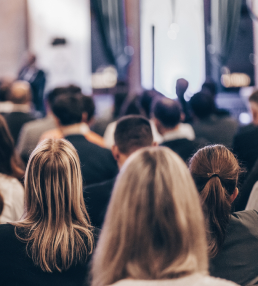 A crowd of people sits in an audience viewing a presentation, with the focus on several attendees' heads and hair from behind while a blurred speaker is visible in the background.