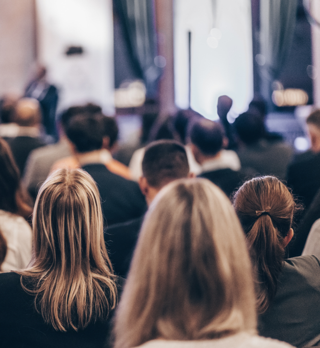 A crowd of people sits in an audience viewing a presentation, with the focus on several attendees' heads and hair from behind while a blurred speaker is visible in the background.
