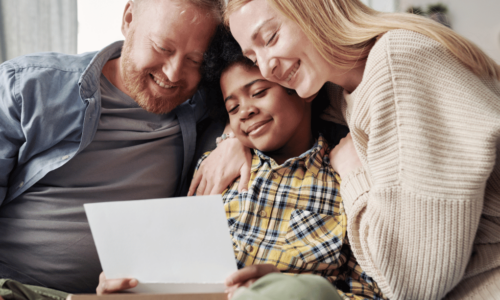 A happy family of three sits close together on a couch, with parents embracing their teenage daughter as they all smile while looking at a document or letter together in a bright, modern living room.