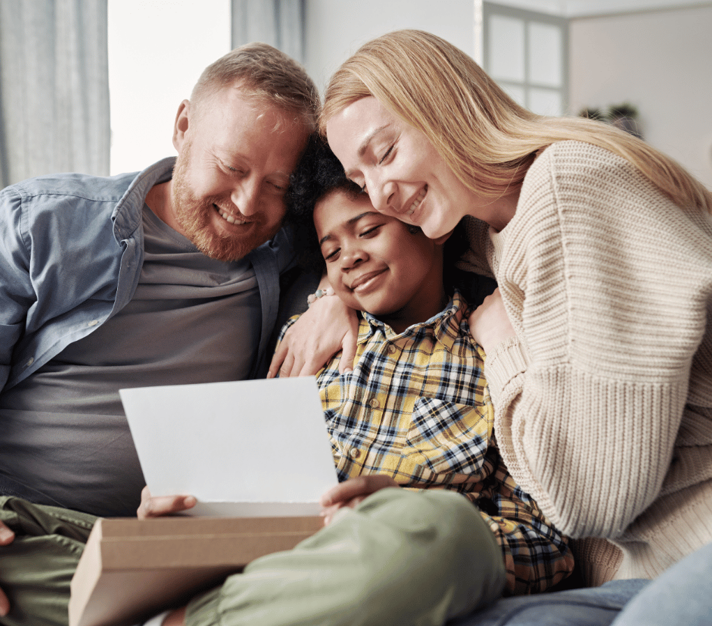 A happy family of three sits close together on a couch, with parents embracing their teenage daughter as they all smile while looking at a document or letter together in a bright, modern living room.