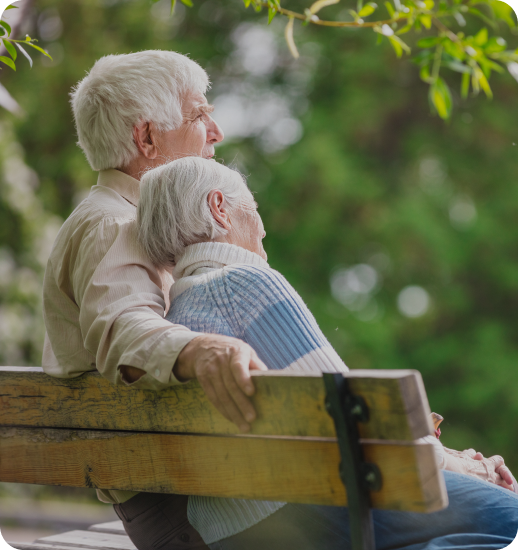 An elderly couple sits together on a wooden park bench, with the man's arm around the woman as they both gaze peacefully into the distance surrounded by lush green foliage.