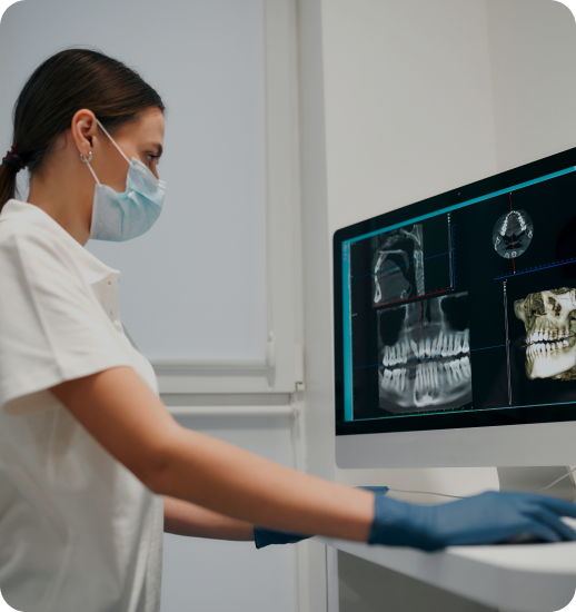 A dental professional wearing a surgical mask and blue gloves examines X-ray images displayed on a computer monitor, showing dental and skull scans in a clinical setting.