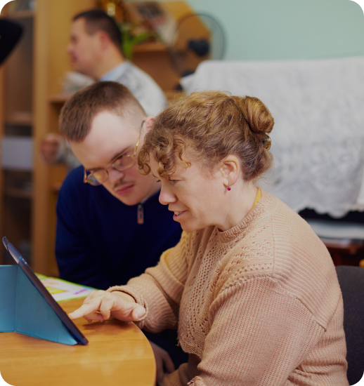 Two people work together at a wooden table, with a woman in a beige sweater and a man in a dark blue shirt looking at documents or a tablet device in what appears to be an office or meeting room setting.