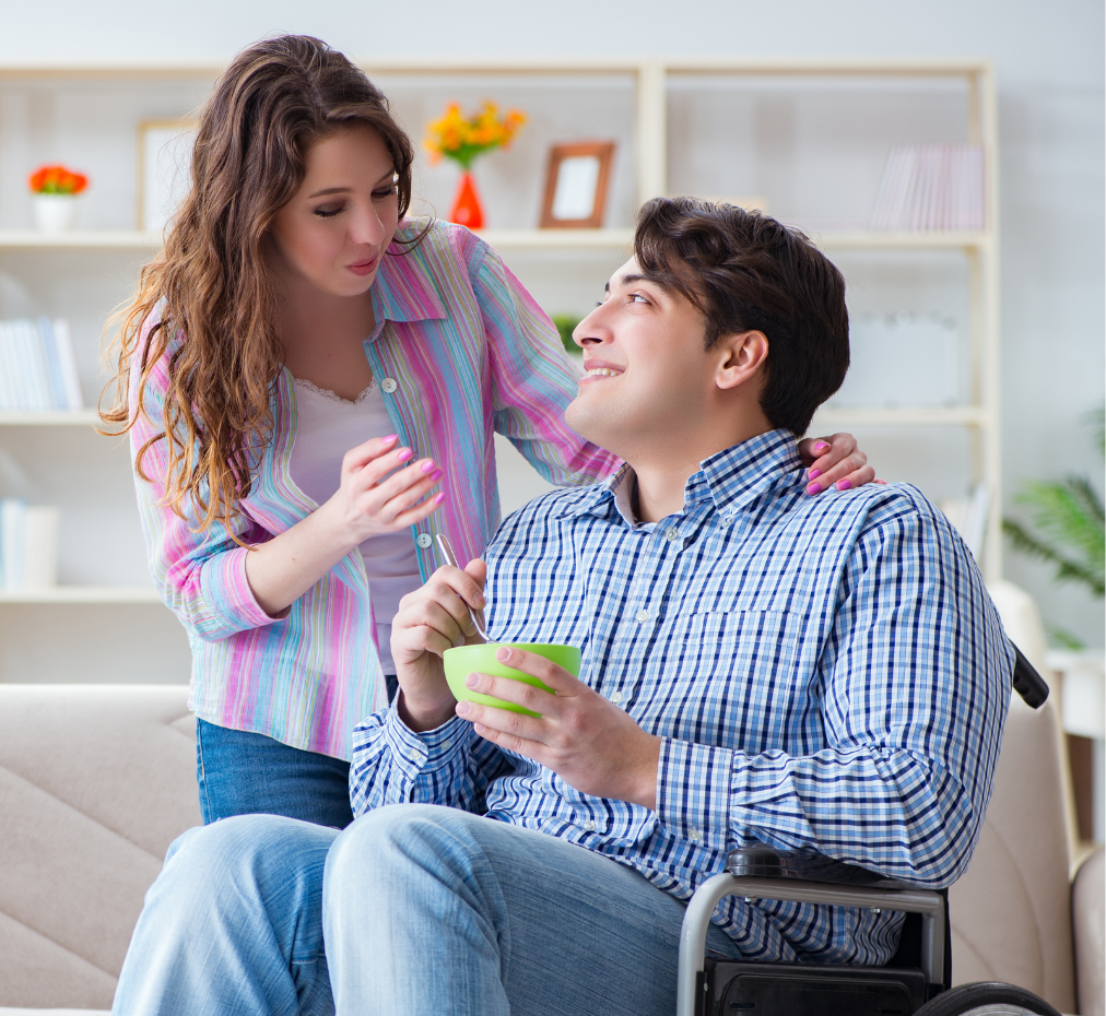 A woman in a colorful striped shirt stands behind a man in a wheelchair wearing a blue checkered shirt, as she helps him eat from a green bowl in what appears to be a home setting with bookshelves in the background.