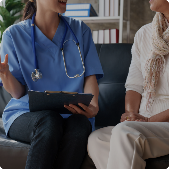 A healthcare professional in blue scrubs with a stethoscope sits on a couch holding a clipboard while speaking with a patient in casual white clothing and a scarf in what appears to be a medical office or consultation room.
