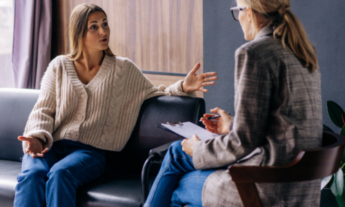 A woman in a cream cardigan sits on a dark couch gesturing with her hands while speaking to a therapist or counselor who is taking notes on a clipboard. The setting appears to be a professional therapy or counseling session in a comfortable office environment with natural lighting.