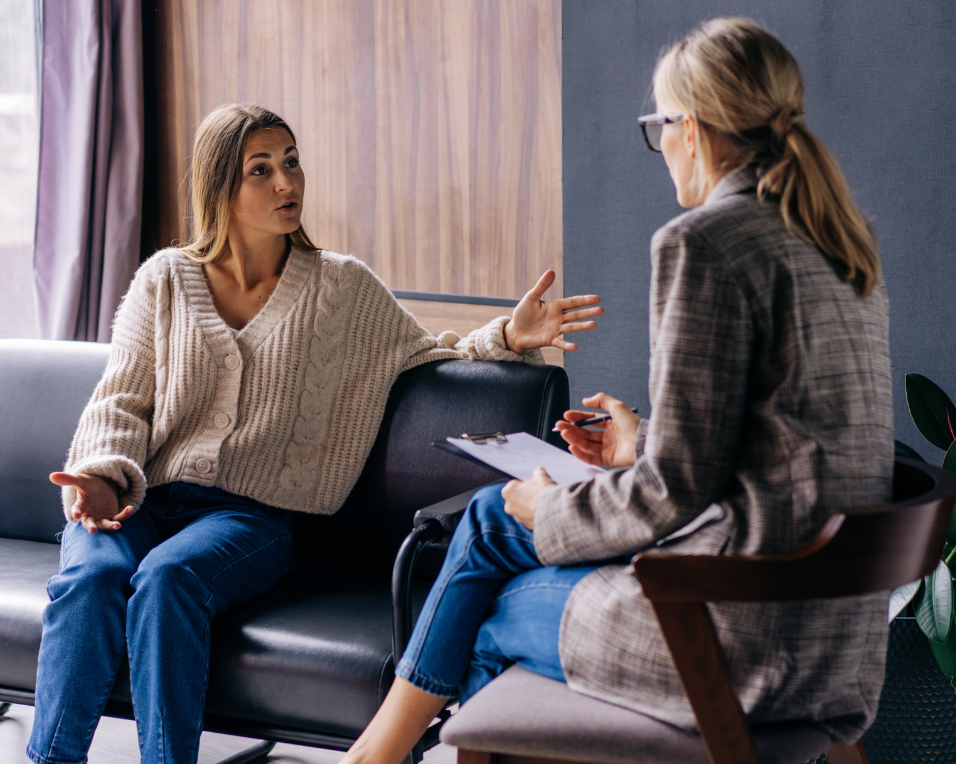 A woman in a cream cardigan sits on a dark couch gesturing with her hands while speaking to a therapist or counselor who is taking notes on a clipboard. The setting appears to be a professional therapy or counseling session in a comfortable office environment with natural lighting.