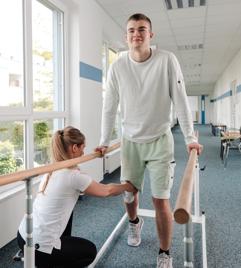 A young man in white shirt and green shorts practices walking with parallel bars while a female physical therapist in white uniform kneels beside him providing support and guidance in a bright rehabilitation facility.