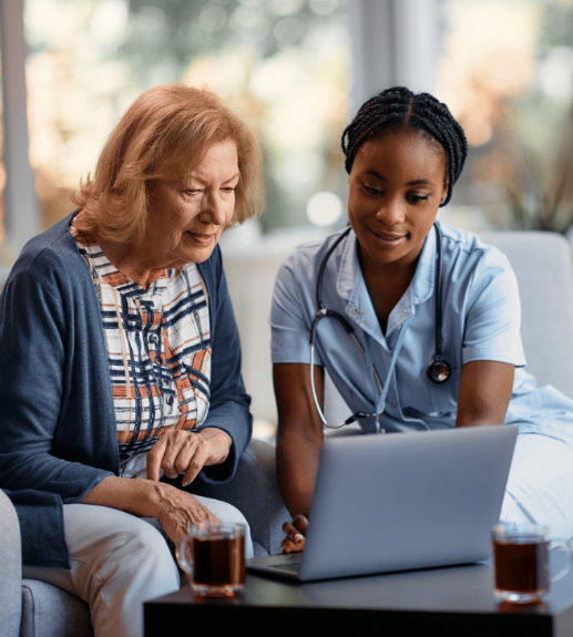 A healthcare professional in scrubs sits with an elderly patient, both looking at a laptop computer together in what appears to be a medical consultation or educational session.