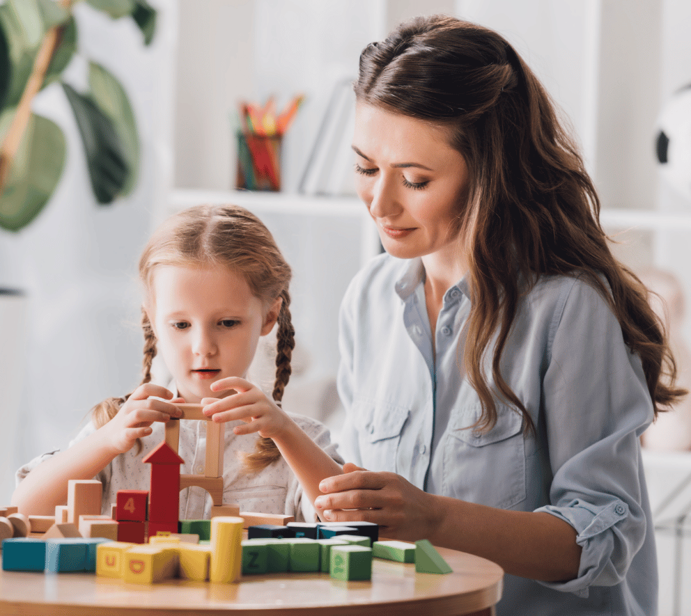 A mother and young daughter sit together at a table, playing with colorful wooden building blocks in a bright, modern living space with white shelving and plants in the background.