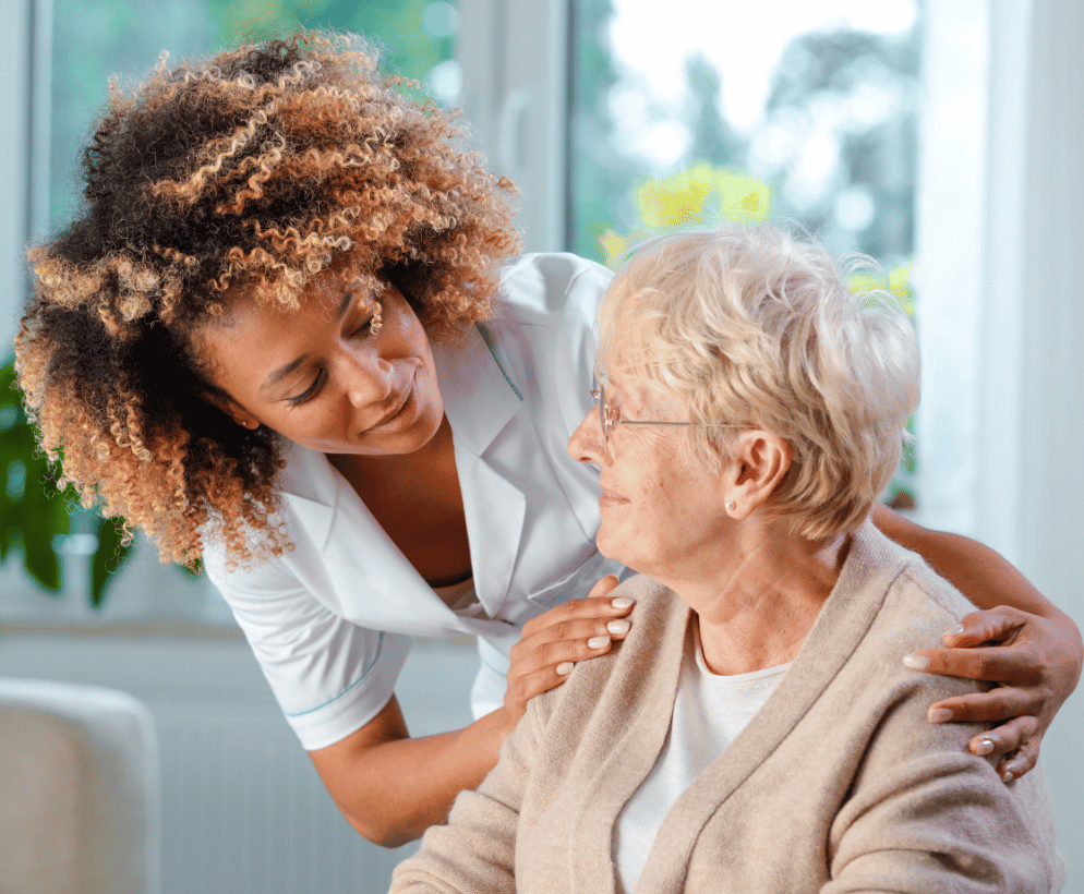 A healthcare worker with curly hair in a white uniform provides compassionate care to an elderly woman with short blonde hair and glasses, gently placing supportive hands on her shoulders in a bright, window-lit room.