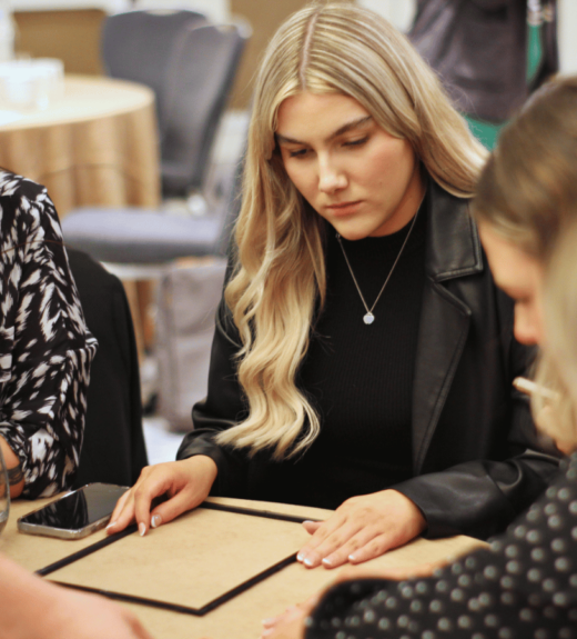 A group of professional women sit around a table in a meeting, with one blonde woman in a black leather jacket looking down at documents while others engage in discussion.