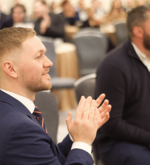 A man in a navy suit and tie sits in the foreground applauding at what appears to be a formal event or conference, with other attendees visible in the background seated in rows of chairs.