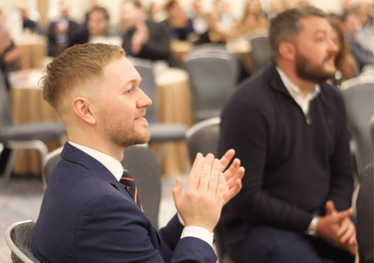 A man in a navy suit and tie sits in the foreground applauding at what appears to be a formal event or conference, with other attendees visible in the background seated in rows of chairs.