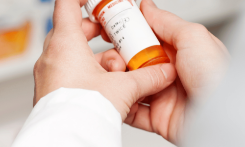 A person's hands holding a prescription medication bottle with an orange and white label, with additional pill bottles visible in the blurred background.
