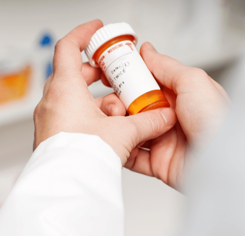 A person's hands holding a prescription medication bottle with an orange and white label, with additional pill bottles visible in the blurred background.