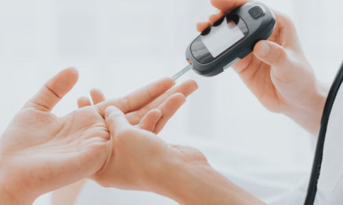 A healthcare professional uses a blood glucose meter to test a patient's blood sugar level by pricking their finger. The device displays the reading on a small digital screen while the patient extends their hand for the test.