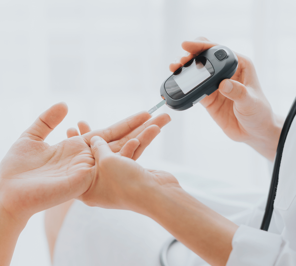A healthcare professional uses a blood glucose meter to test a patient's blood sugar level by pricking their finger. The device displays the reading on a small digital screen while the patient extends their hand for the test.