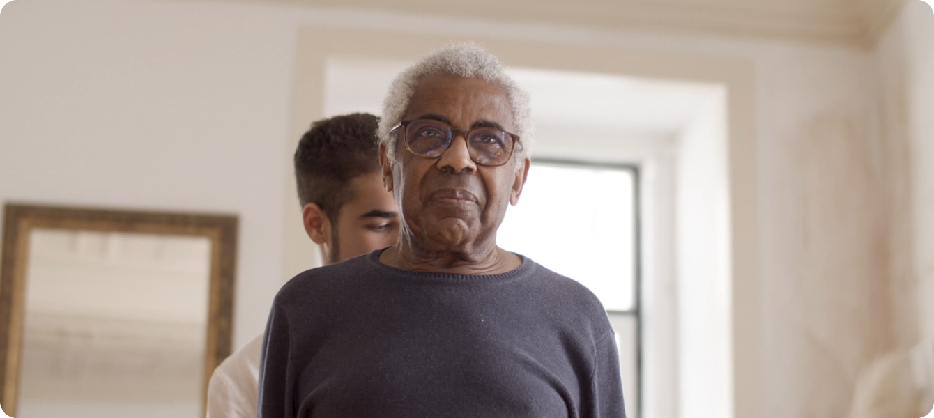 An elderly Black man with white hair and glasses wearing a dark sweater stands in the foreground of a bright, modern home interior, with a young person visible behind him near a window.