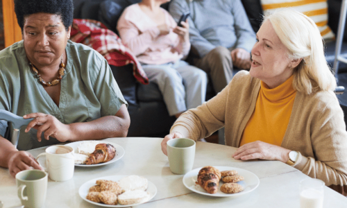 Two women sit at a white table having coffee and pastries, with one woman wearing a yellow turtleneck and cardigan speaking to another in a light green shirt. In the background, an elderly couple sits together on a dark couch in what appears to be a comfortable living room setting.
