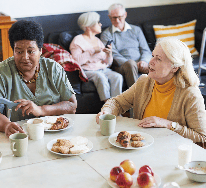 Two women sit at a white table having coffee and pastries, with one woman wearing a yellow turtleneck and cardigan speaking to another in a light green shirt. In the background, an elderly couple sits together on a dark couch in what appears to be a comfortable living room setting.
