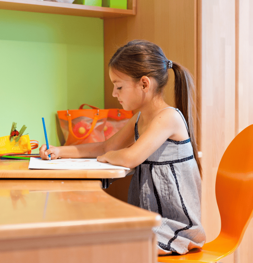 A young girl with her hair in a ponytail sits at a wooden desk writing or drawing on paper with a blue pen, surrounded by colorful school supplies in what appears to be a bright, modern classroom or study area.