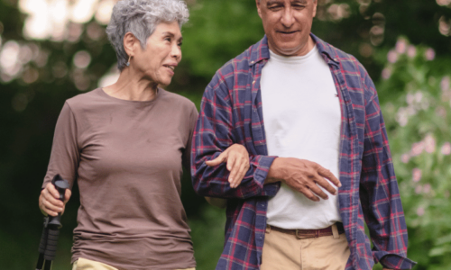 An elderly couple walks together outdoors, with the woman holding a walking stick and linking arms with the man who wears a plaid shirt, both smiling as they stroll through a green, tree-lined area.